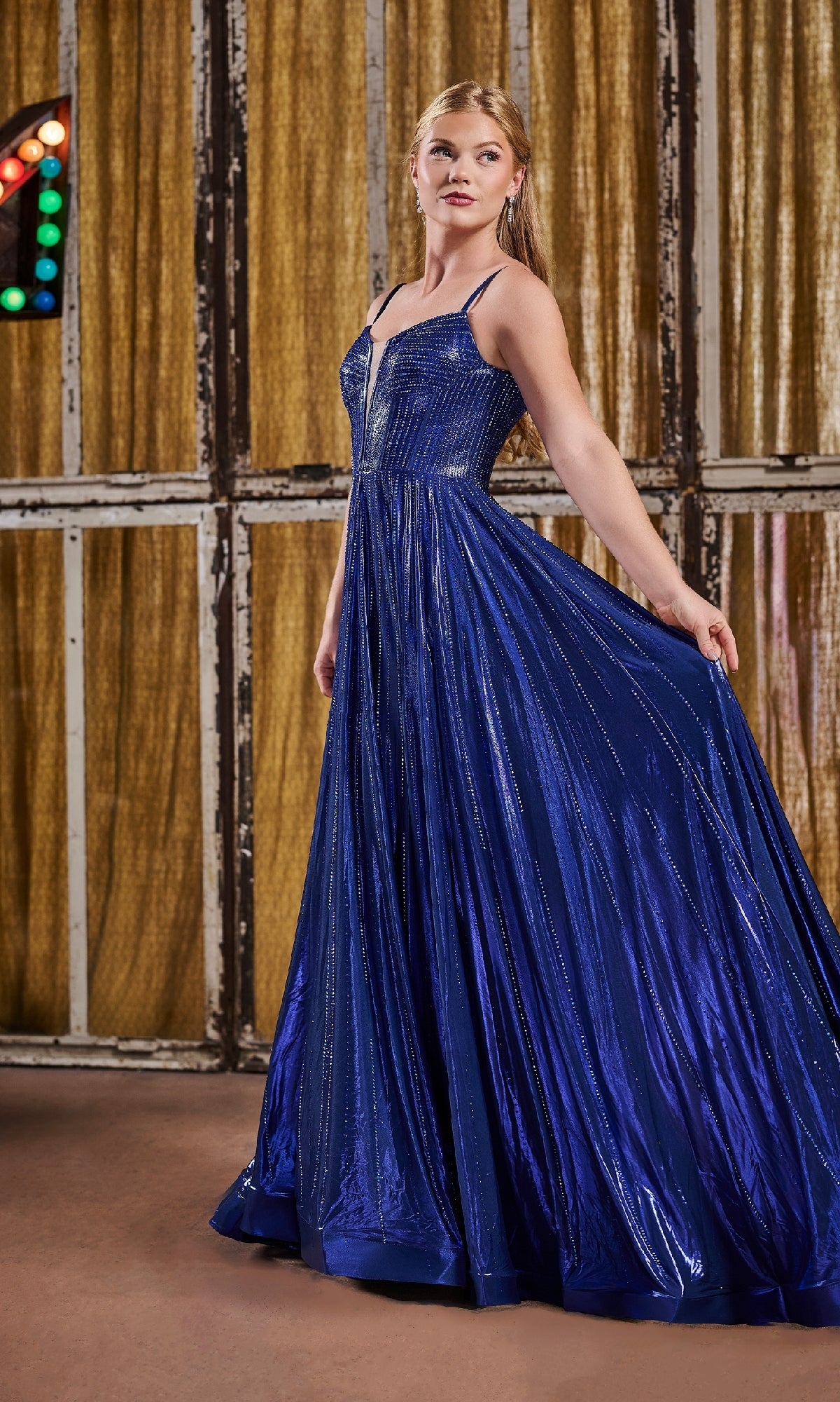 A woman stands indoors holding out one side of her floor-length, sleeveless blue Tiffany 16189 Long Prom Dress; gold curtains and multicolored lights decorate the background.